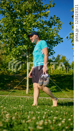Older man walking barefoot on grass holding sneakers under bright sunlight 128828121