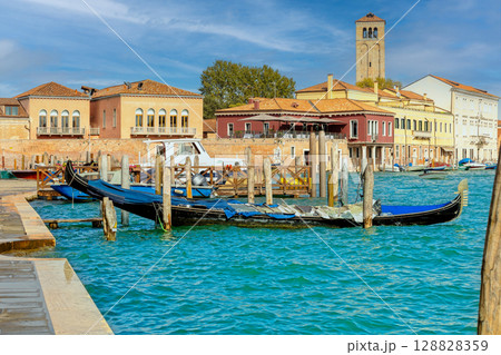 Traditional Venetian waterfront scene on Murano island with gondolas, colorful buildings, and a historic bell tower under sunny skies. Traditional Venetian waterfront scene on Murano island with gondolas, colorful buildings, and a historic bell tower under sunny skies. 128828359