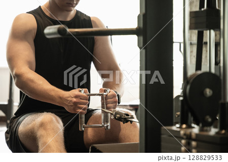 Young caucasian man wearing blank sleeveless tank top exercising on rowing machine at gym. Close up. 128829533