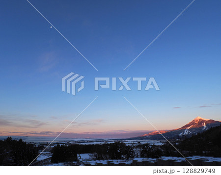 冬の早朝、朝日が当たり、赤く輝く冠雪の磐梯山。青空に白く輝く月が印象的で綺麗だ。 128829749