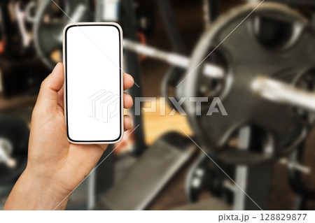 Mans hand holding smartphone with white blank screen in blurred gym with bench press and weight plates as background. Workout plan. Mock up. Mans hand holding smartphone with white blank screen in blurred gym with bench press and weight plates as background. Workout plan. Mock up. 128829877