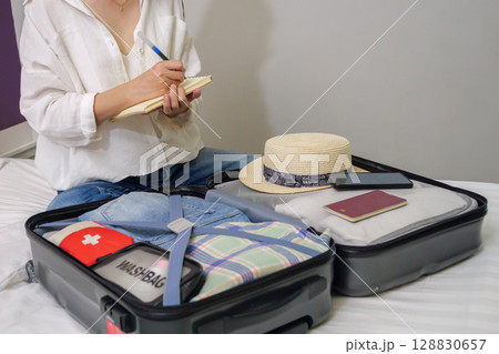 making a check list of things to pack for travel. woman writing checklist item on notebook and packing suitcase to vacation, prepare clothes, medicine, passport into luggage. travel vacation concept making a check list of things to pack for travel. woman writing checklist item on notebook and packing suitcase to vacation, prepare clothes, medicine, passport into luggage. travel vacation concept 128830657