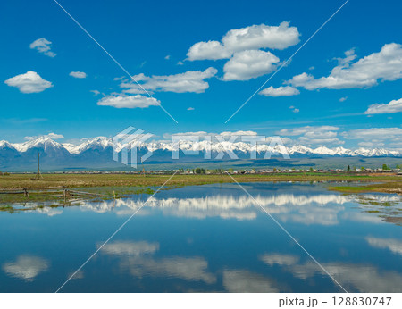Majestic snow-capped mountains reflect in serene lake under bright blue sky with fluffy clouds in summer 128830747