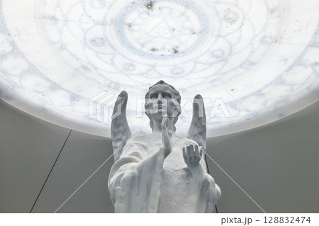 Angelic white statue praying beneath ornate luminous dome in modern sacred interior 128832474