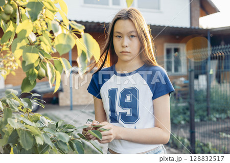 serious teenage girl in sporty t shirt standing by green tree branches in backyard garden in front of house during sunset in summer concept of natural childhood growth, rural outdoor 128832517