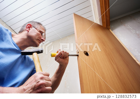 Creating precise mortise with chisel and hammer, installer shapes cavity inside door edge to fit lock mechanism as part of interior door renovation. 128832573