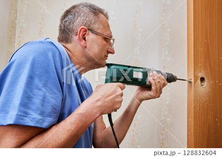 Drilling hole for latch bolt with spade bit in wooden door edge using electric drill during lock installation process. 128832604