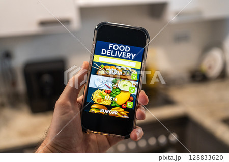 Male hands holding phone with food delivery app on screen against background of room in house 128833620
