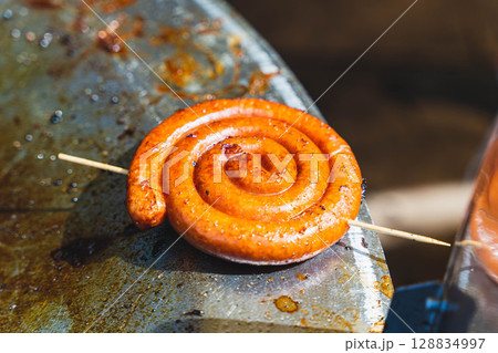 Grilled spiral sausage cooking on a hot surface with wooden skewer Grilled spiral sausage cooking on a hot surface with wooden skewer 128834997