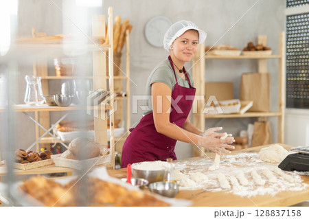 Female baker prepares raw dough in bakery, forming it into long sausages to be cut into pieces for baking buns or croissants 128837158