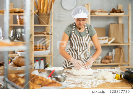 Young smiling female baker preparing raw yeast dough on kitchen table in bakery 128837789