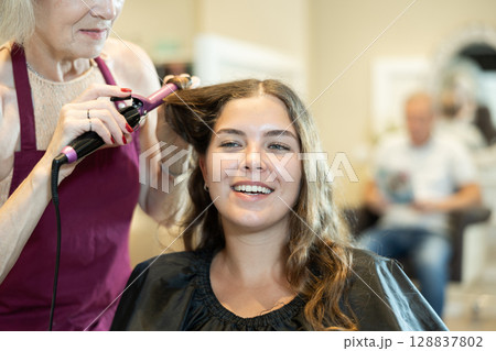 In beauty salon, cropped elderly female hairdresser makes styling curls for young girl client In beauty salon, cropped elderly female hairdresser makes styling curls for young girl client 128837802