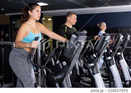 Athletic young woman exercising on an elliptical trainer in gym Athletic young woman exercising on an elliptical trainer in gym 128837859
