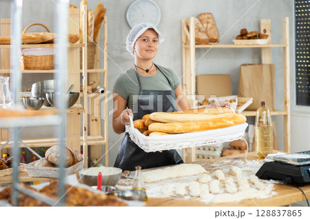 Bakery female seller stands proudly in front of fresh bread holding basket of freshly baked baguettes in kitchen 128837865