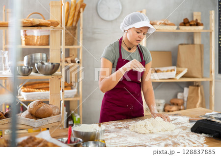 Woman works in bakery as baker, kneads dough, works with flour. 128837885