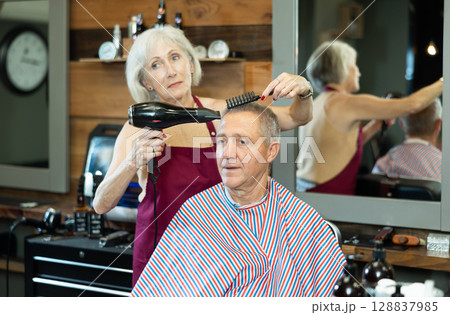 Female barber using hairdryer to style hair to elderly client in barbershop 128837985