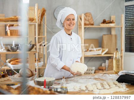 Young baker girl is standing near work table in kitchen, working with dough Young baker girl is standing near work table in kitchen, working with dough 128837997