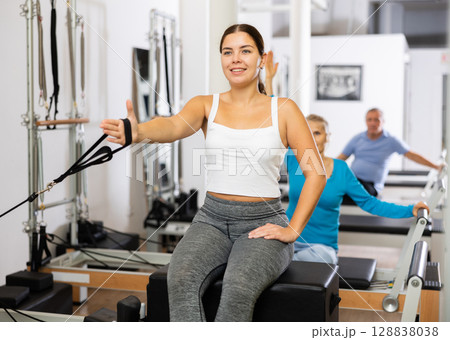Young woman in sportswear sitting and doing Pilates exercises for hands on reformer bed equipment in fitness center 128838038