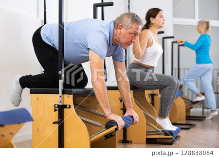 Focused man and women of different ages performing stretching exercises on Wanda Chair equipment in Pilates studio Focused man and women of different ages performing stretching exercises on Wanda Chair equipment in Pilates studio 128838084