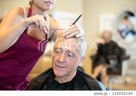 Cropped young girl hairdresser performs haircut for elderly man using scissors Cropped young girl hairdresser performs haircut for elderly man using scissors 128838104