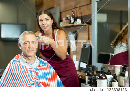 Young female barber making haircut to elderly client in barbershop 128838138