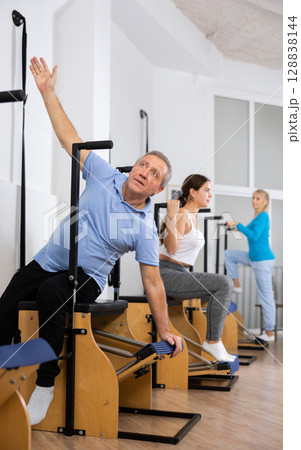 Elderly male in activewear sitting and training on Wanda chair machine during Pilates class Elderly male in activewear sitting and training on Wanda chair machine during Pilates class 128838144