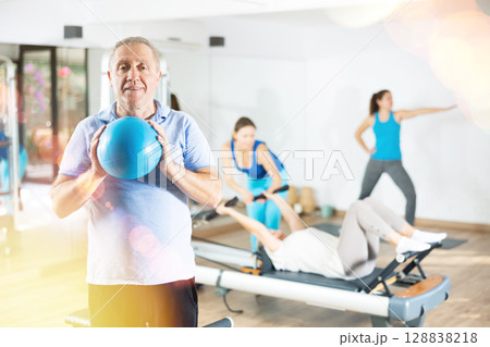 Smiling mature male squeezing ball in hands during Pilates training in gym Smiling mature male squeezing ball in hands during Pilates training in gym 128838218