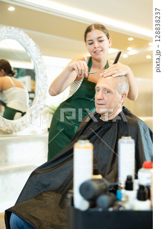 Young girl hairstylist making haircut to elderly man in hairdressing salon 128838237