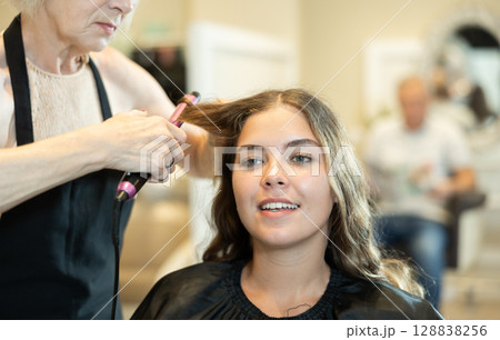 Female hairdresser makes curls the hair of young woman using curling iron Female hairdresser makes curls the hair of young woman using curling iron 128838256