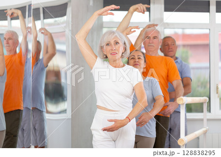 Active mature woman practicing third position holding by ballet barre together with others 128838295