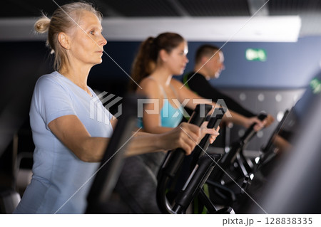 Sporty senior woman exercising on elliptical trainer in gym Sporty senior woman exercising on elliptical trainer in gym 128838335