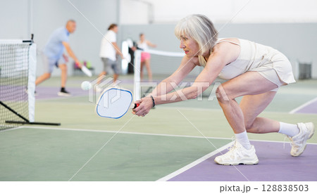 Elderly woman bouncing ball in pickleball game on court with racket 128838503