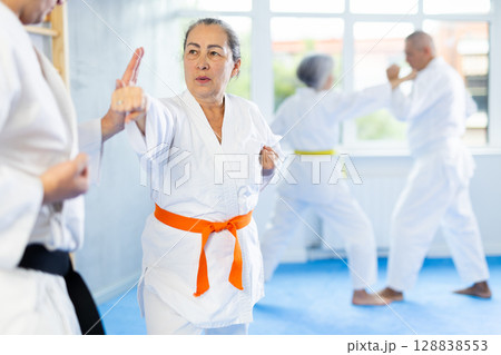 Old woman attendee of karate classes fighting with her opponent in sports hall Old woman attendee of karate classes fighting with her opponent in sports hall 128838553