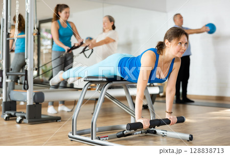 Peaceful middle-aged female in activewear concentrating on stretching exercise on Pilates chair equipment in gym Peaceful middle-aged female in activewear concentrating on stretching exercise on Pilates chair equipment in gym 128838713
