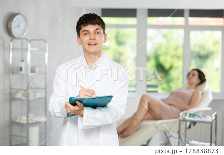 Smiling man beautician in uniform standing in cosmetology clinic, filling out clipboard with medical records Smiling man beautician in uniform standing in cosmetology clinic, filling out clipboard with medical records 128838873