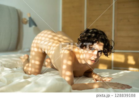Fully naked woman posing erotically on her knees in the bedroom on the bed in the light of the sun's rays breaking through window blinds 128839108
