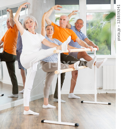 Dedicated mature females and males ballet dancer performing leg stretch at barre with concentration and grace in choreography studio during group rehearsal 128839241