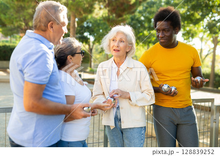 Mature multiracial casual friends smiling while holding metal petanque balls outdoors 128839252