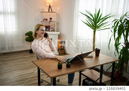 Smiling woman engaged in a phone conversation while working from a cozy home office with plants and natural light 128839401