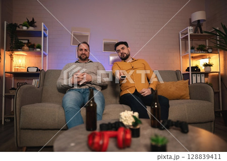 Two friends enjoying a gaming night together in a cozy, well-lit living room with drinks and controllers at hand 128839411