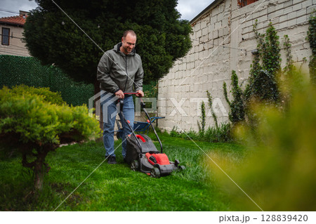 Man mowing a lush green lawn in a serene backyard garden during a cloudy afternoon 128839420
