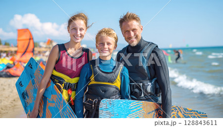 Smiling family dressed in wetsuits, life vests, stands on sunny beach holding kitesurfing boards. Ocean stretches behind them, with kitesurfers in distance, scene of adventure, shared passion 128843611