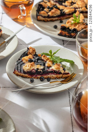 A slice of homemade blueberry crumble pie on a plate on white tile background. Sweet breakfast composition in bright morning light 128844296
