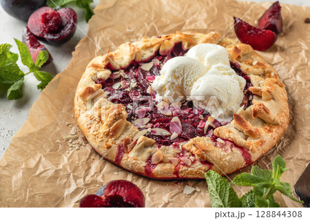 French Galette or pie with plums and almonds served with ice cream scoops on parchment paper background. Summer traditional homemade sweet pastries 128844308