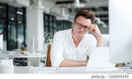 Man wearing glasses, white shirt sits at desk in modern office, looking tired and rubbing his forehead while looking at laptop. Themes of stress, fatigue, challenges of demanding work environment. 128844340