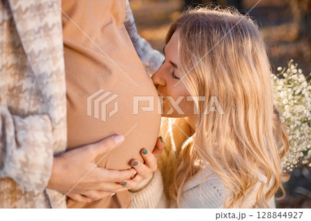 Close-up view of happy girl near a pregnant belly. Women expecting a baby and standing in a autumn park. Portrait of curly blonde woman. 128844927