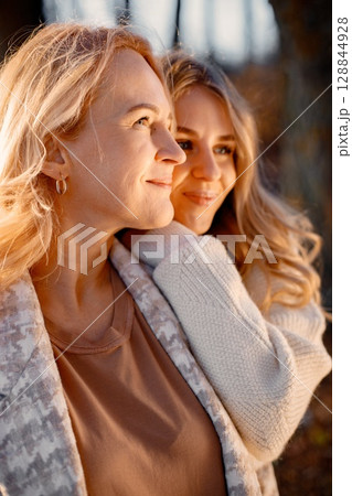 Mature mother with her teenage daughter hugging in a park. Women looking in one direction. Blonde women wearing beige clothes. Mature mother with her teenage daughter hugging in a park. Women looking in one direction. Blonde women wearing beige clothes. 128844928