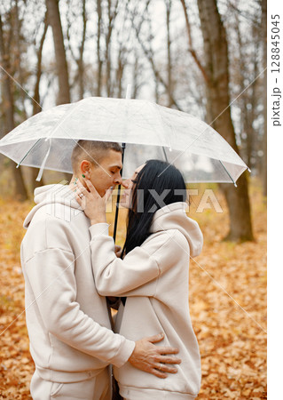 Man and woman standing in autumn forest and kissing under transparent umbrella. Brunette man and woman falling in love. Family wearing beige sportive costumes. 128845045