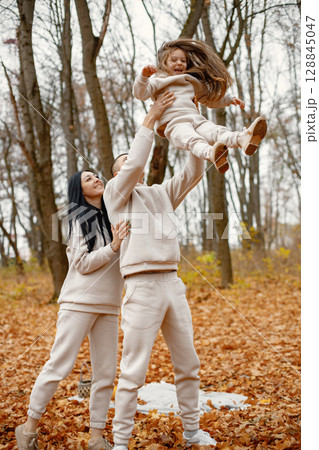 Man, woman and their little girl standing in autumn forest and laughing. Brunette man throwing his daughter in the air. Family wearing beige sportive costumes. 128845047