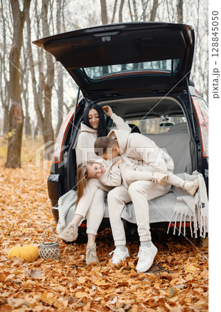 Family resting after day spending outdoor in autumn park. Father, mother, their daughter hugging inside car trunk, smiling. Family wearing beige sportive costumes. 128845050